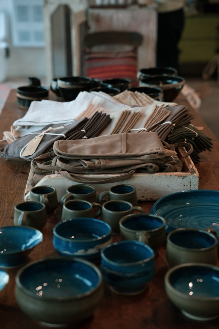 Handmade blue and green glazed bowls and mugs with folded linen napkins and aprons on a wooden table at the Cabin